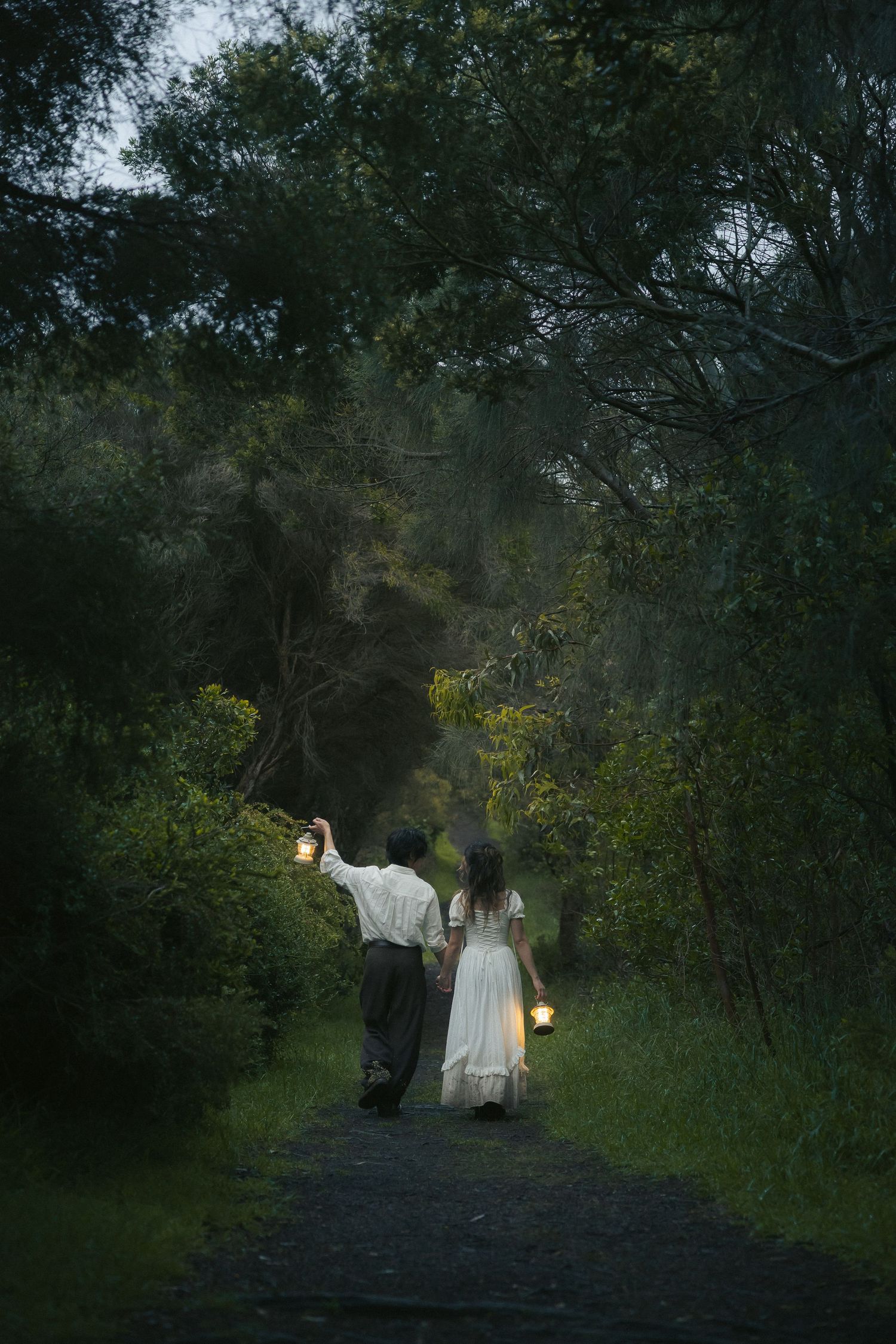 A couple in vintage clothing walks down a dark forest path holding lanterns, creating a mystical evening atmosphere.