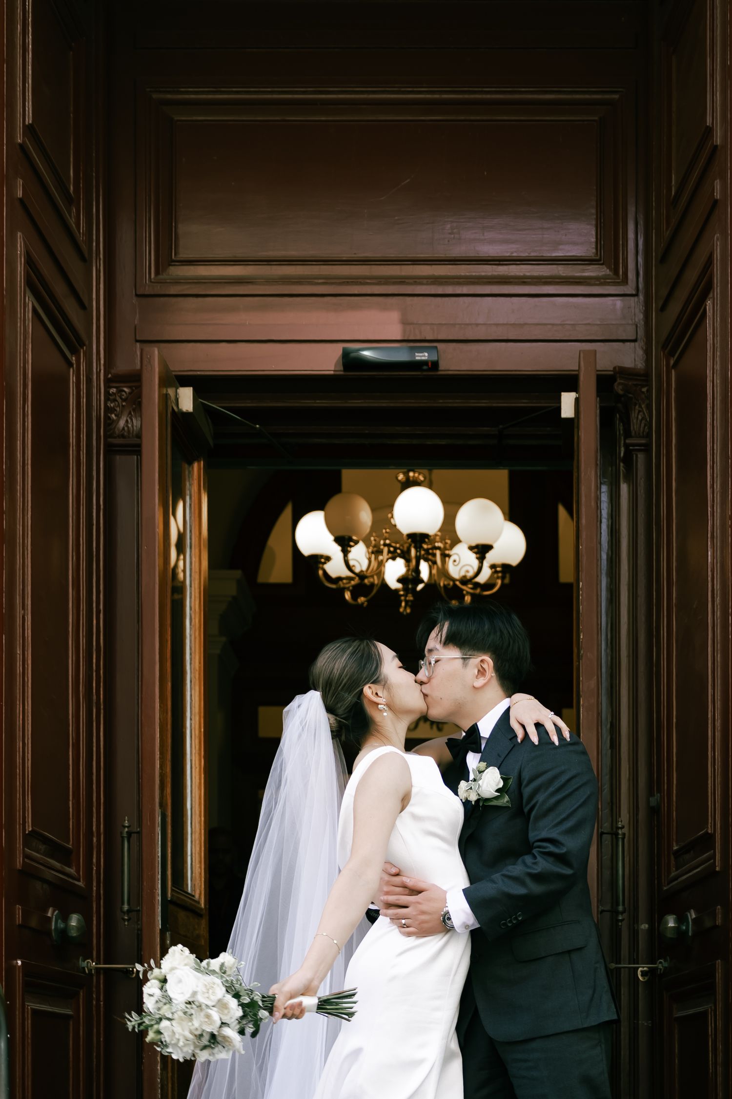A romantic wedding couple shares a tender kiss in an elegant wood-paneled hallway with ornate lighting fixtures.