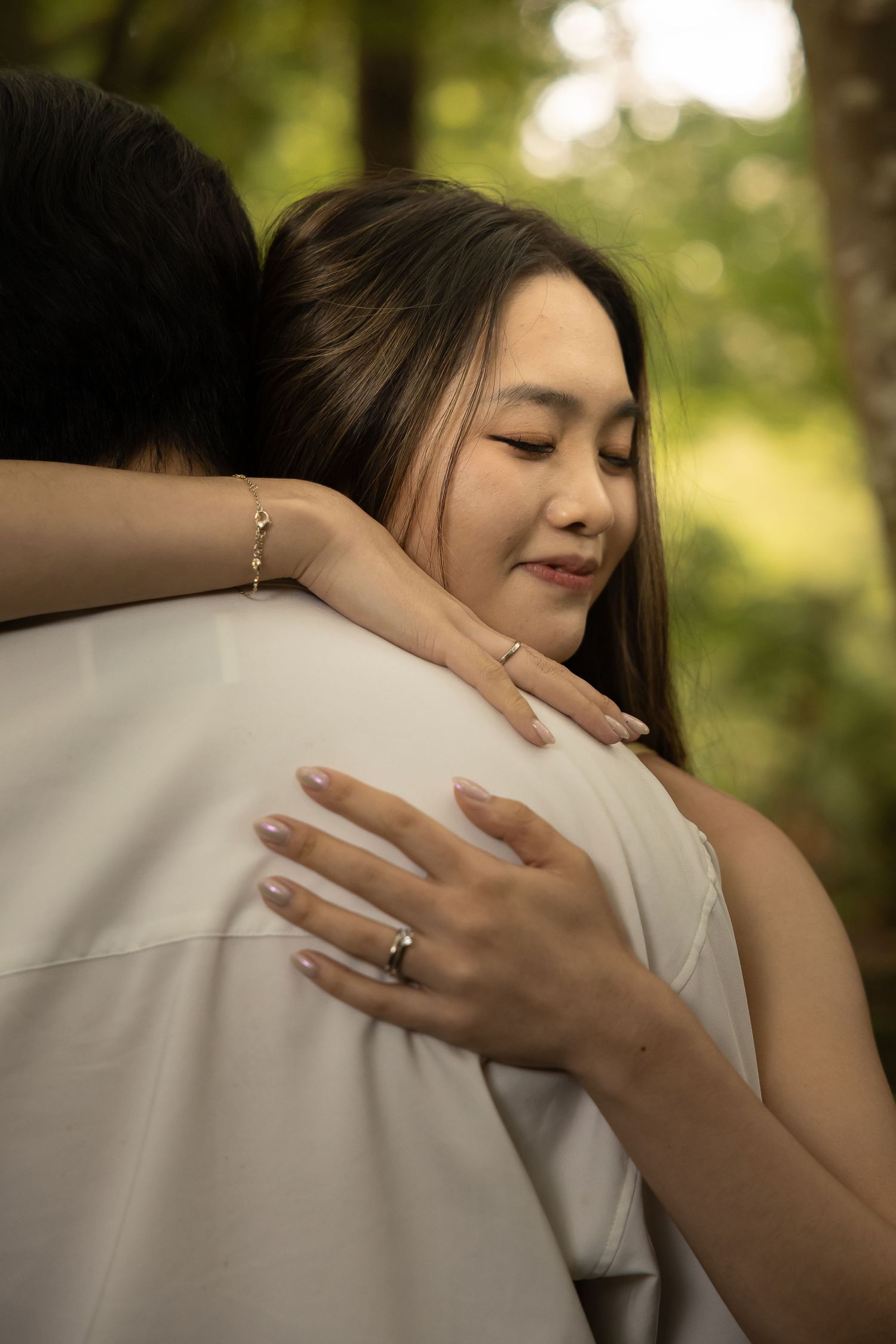 A tender embrace shows hands with an engagement ring resting against a white shirt in soft natural lighting.