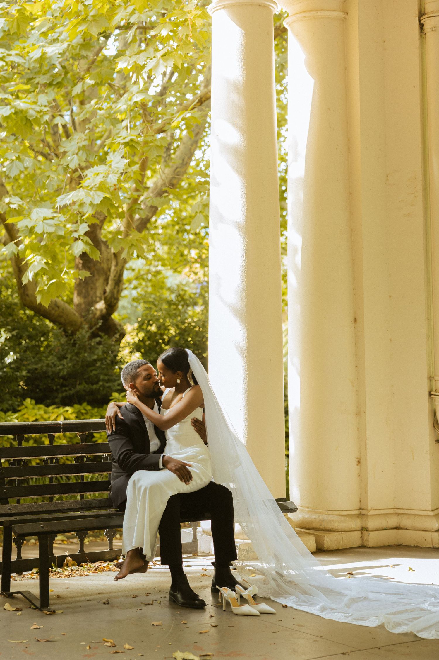 A romantic wedding couple shares intimate moments on a bench surrounded by elegant white columns in a garden setting.