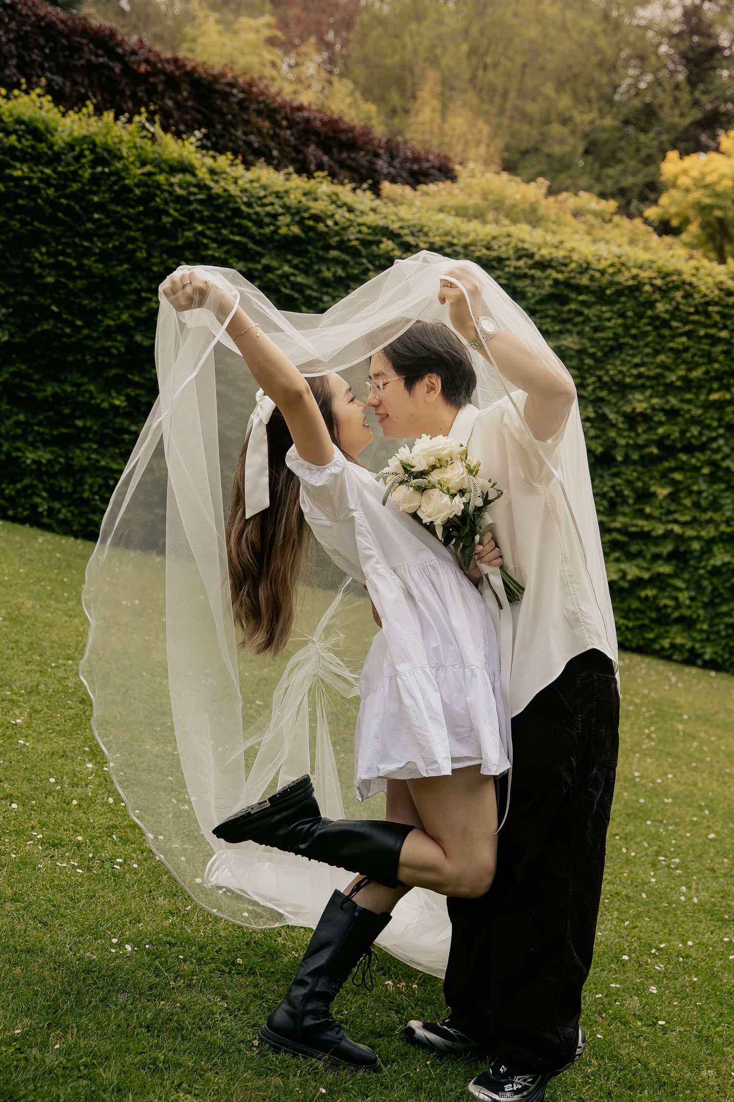 A playful couple shares a moment under a flowing wedding veil in a garden setting.