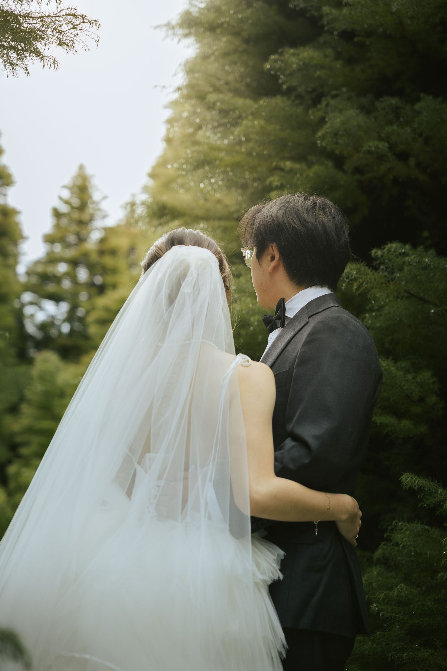 A couple in wedding attire embrace outdoors against a backdrop of evergreen trees in a romantic garden setting.