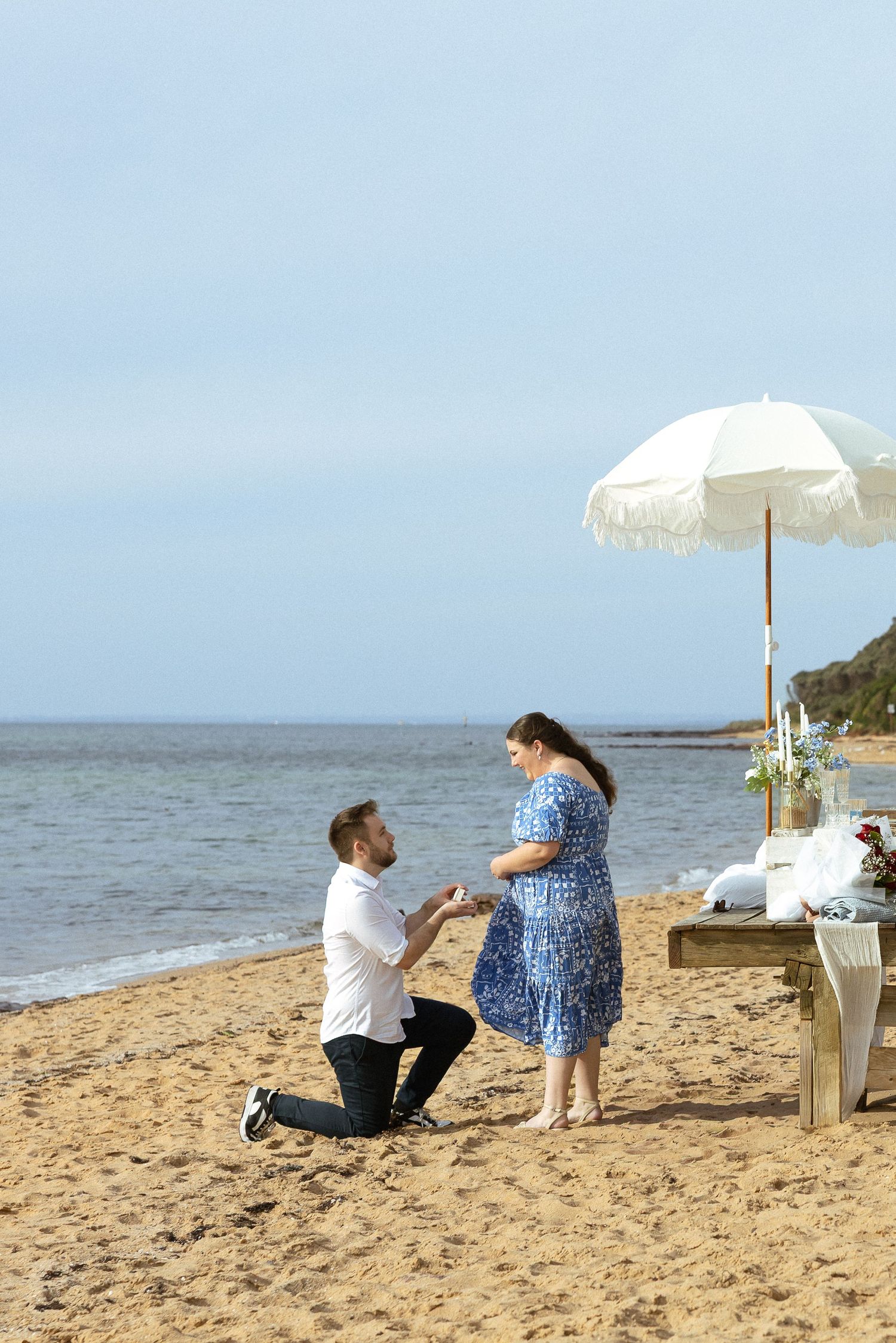 A romantic beach proposal scene unfolds as a couple shares a special moment on the sandy shore with a white umbrella nearby.