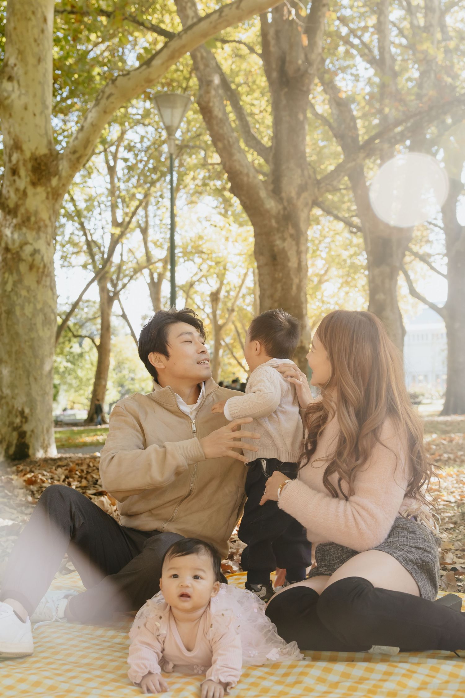 A family sits together on a picnic blanket in a sunlit park during autumn with fallen leaves around them.