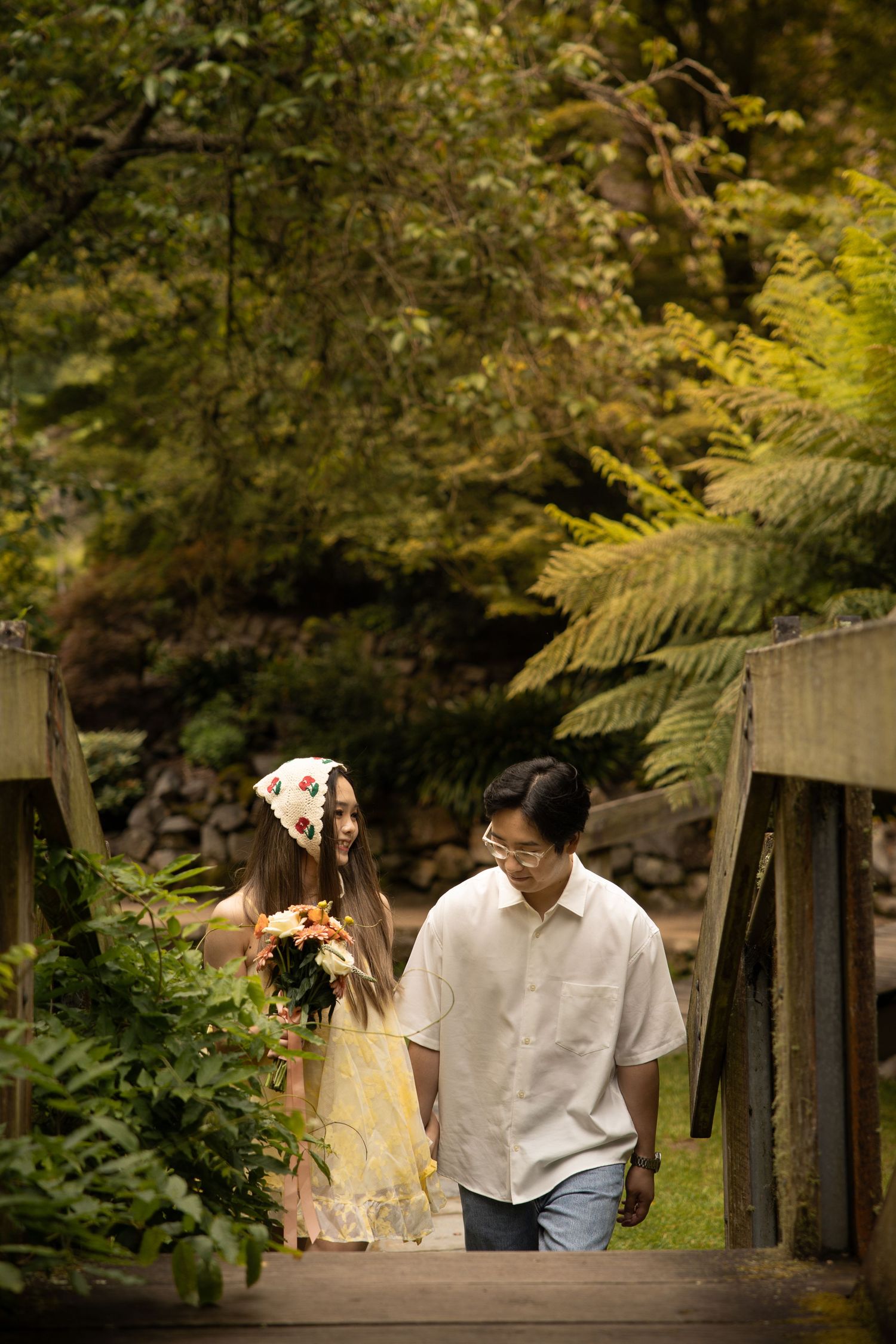 A couple shares a tender moment while walking across a wooden bridge in a serene garden setting.