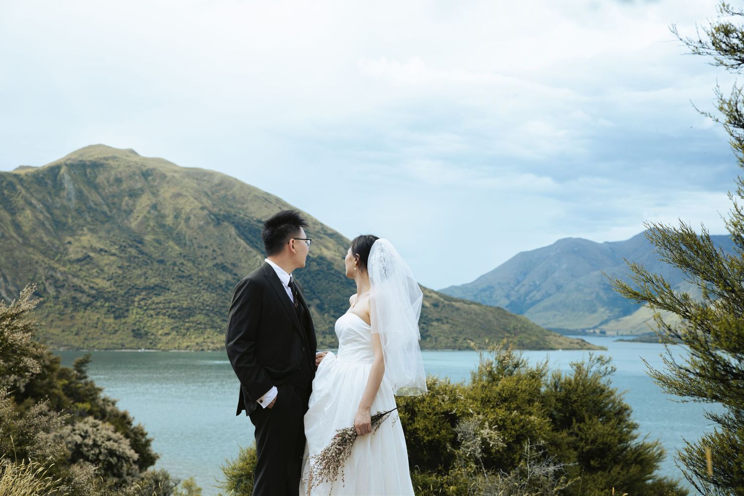 A couple in wedding attire stands on a scenic mountain overlook with a lake and mountains in the background.