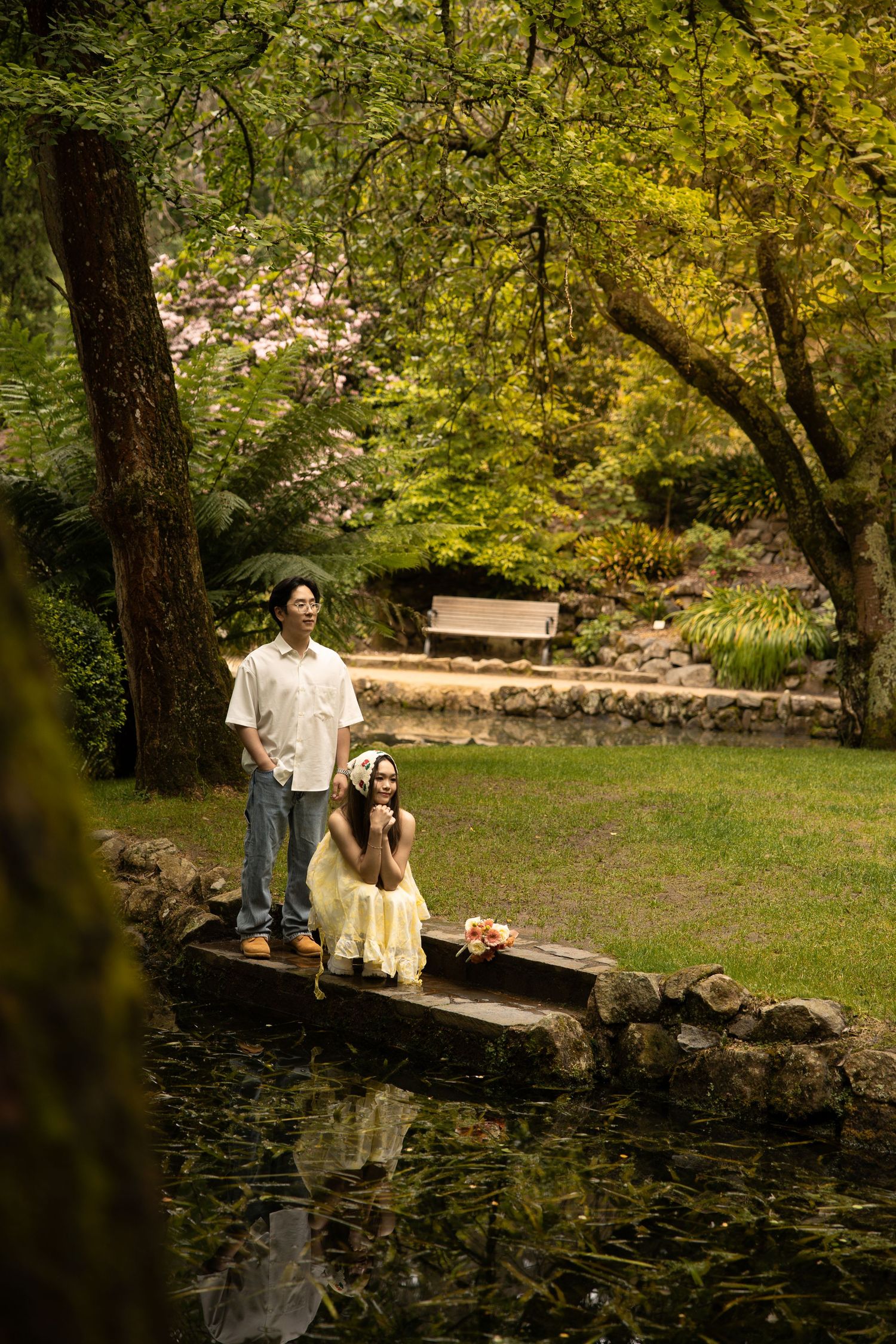 Two people stand on a wooden bridge over a reflective pond in a lush garden setting with green trees and foliage.