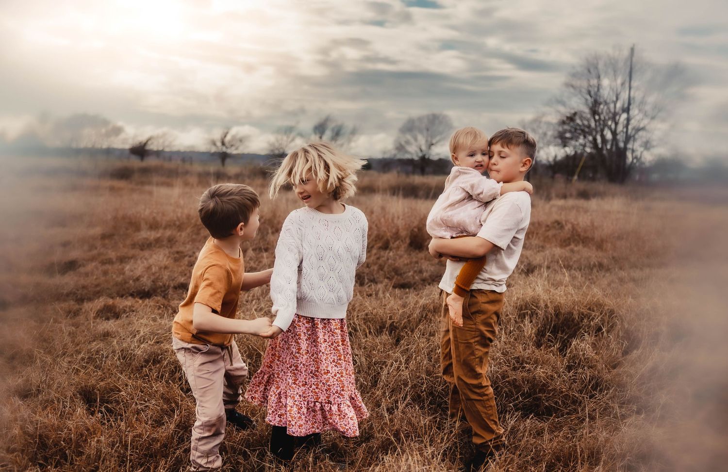 Three children stand together in a brown autumn field at golden hour under a moody cloudy sky.