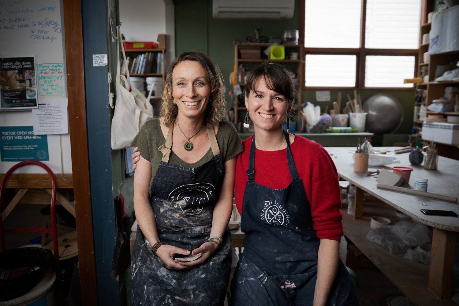 Two smiling women wearing aprons posing together in a pottery studio workshop.