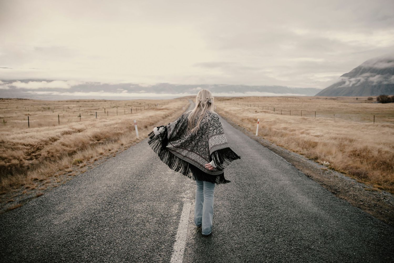Woman in patterned wrap walking down empty rural road with mountains ahead.
