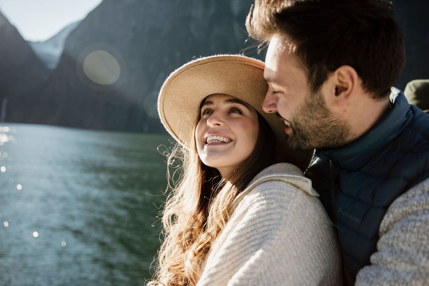 Smiling couple embracing near a sunlit mountain lake on a bright day.
