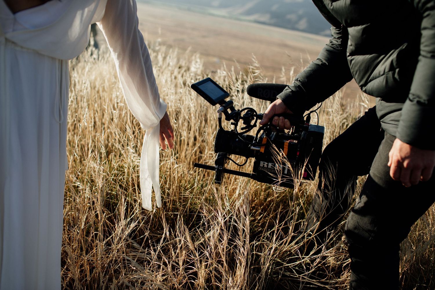 Videographer filming woman in white dress among golden grass fields with professional camera rig outdoors.