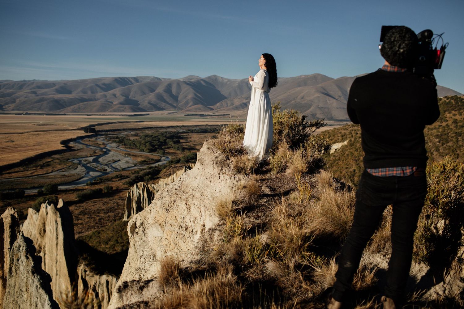 Photographer capturing a woman in white dress on rocky cliff with scenic mountain valley backdrop in New Zealand.