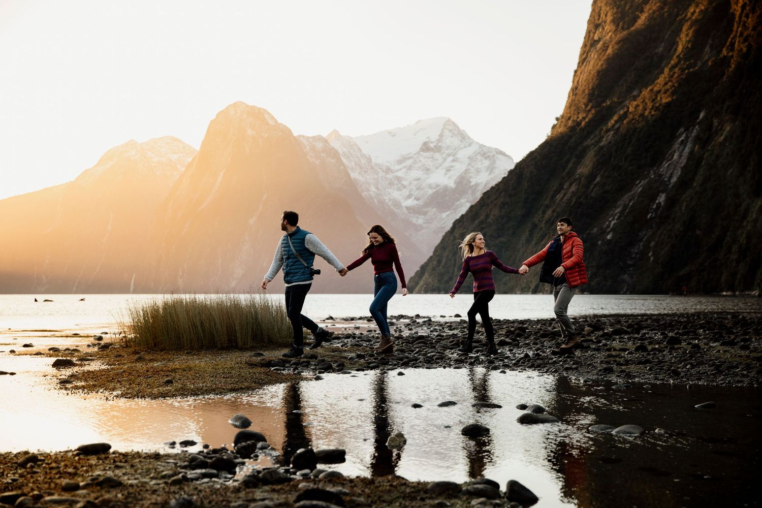 Four people holding hands walking on rocky shore at golden hour near mountains.