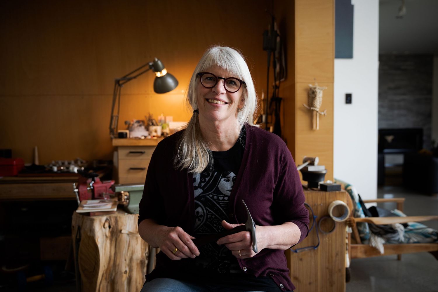 Smiling senior woman artist sitting in creative workshop studio surrounded by tools and artwork.