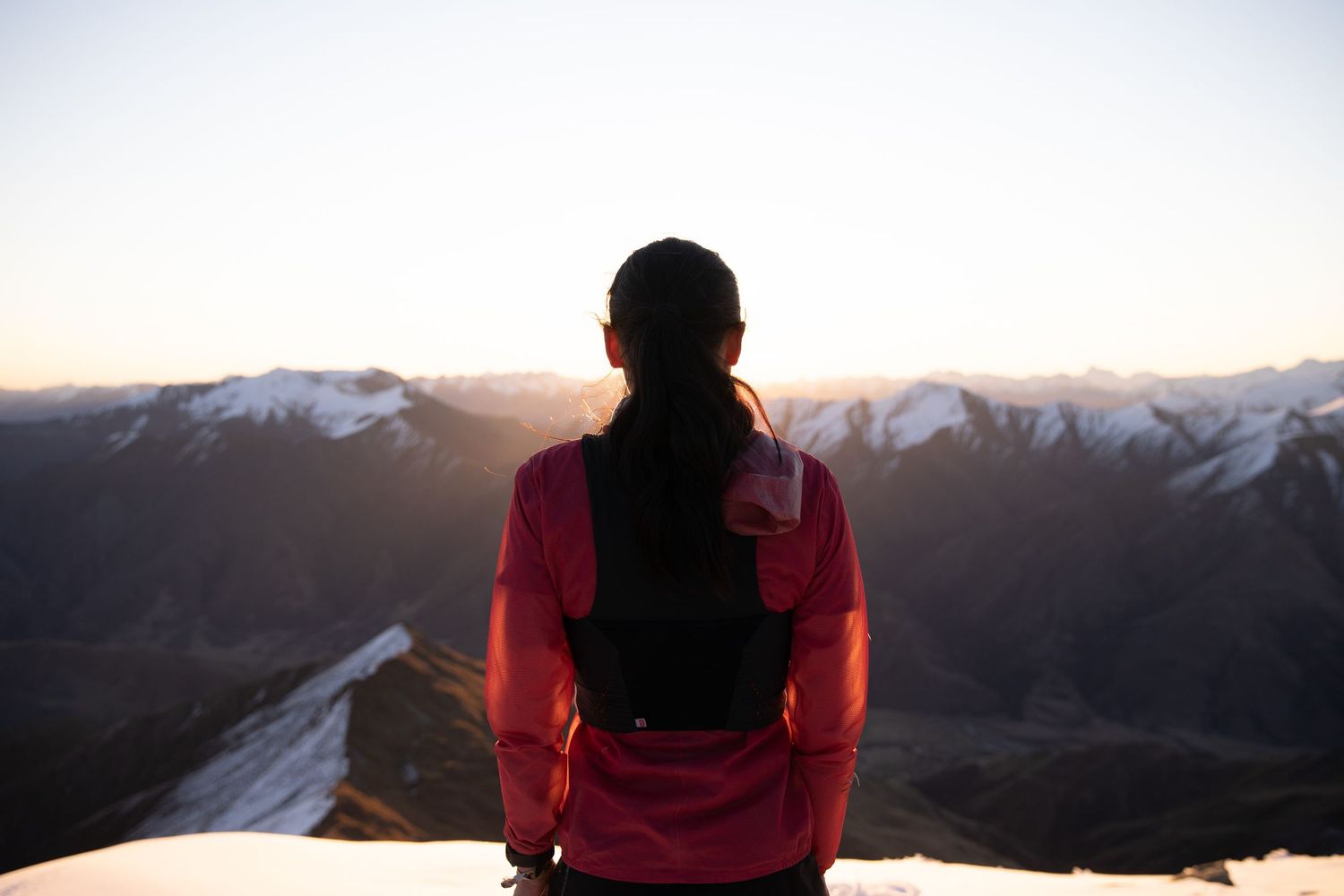 Hiker in red jacket stands on snowy mountain summit admiring panoramic views at sunrise.