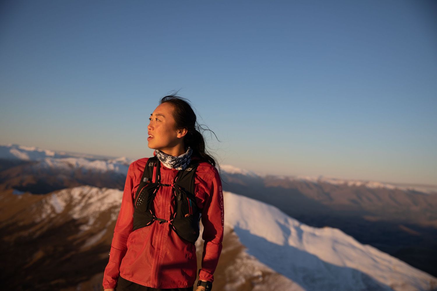 Female hiker in red jacket stands on snowy mountain peak during golden hour sunset.