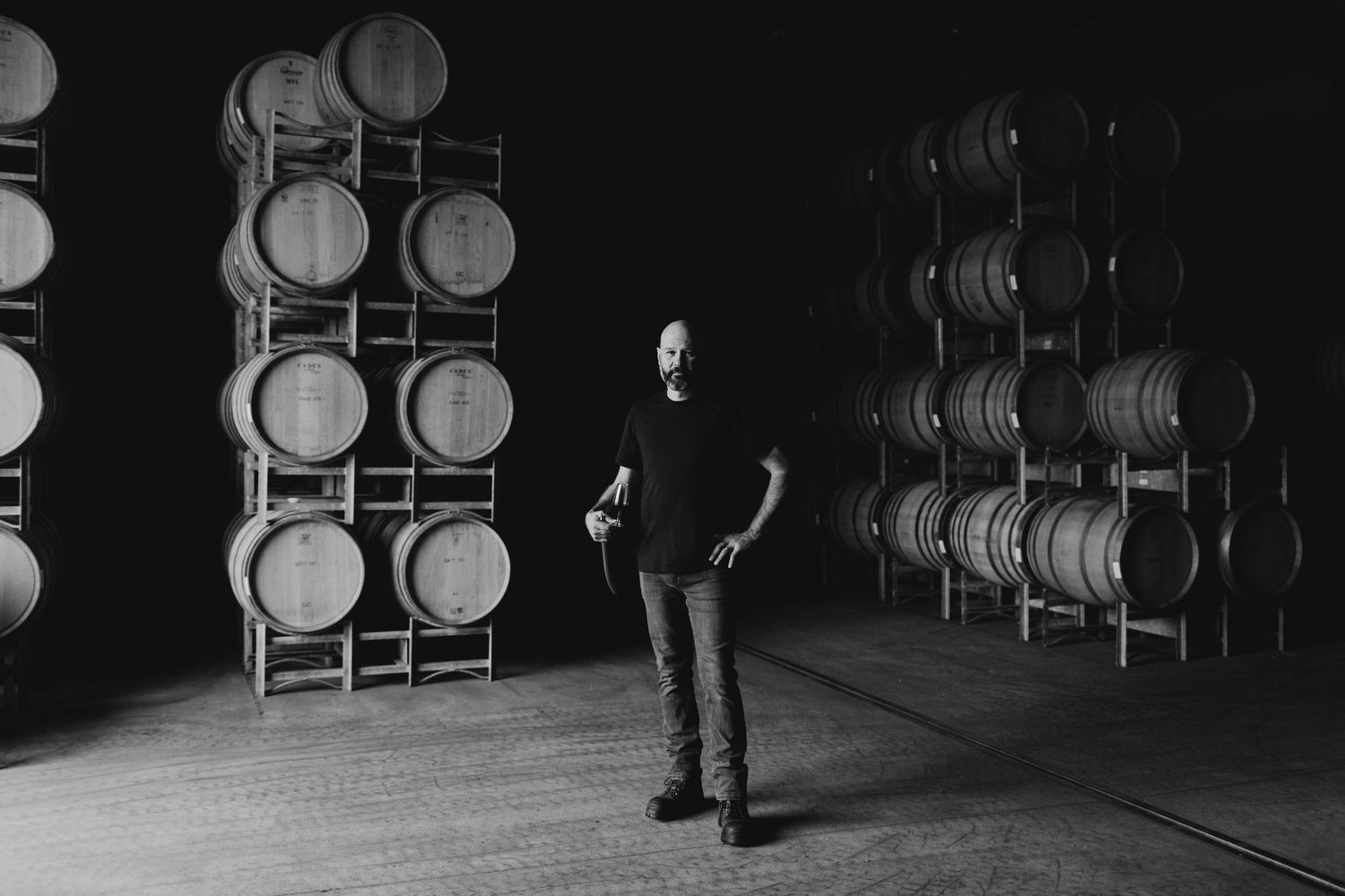 Man standing in dimly lit winery barrel room surrounded by oak wine barrels stacked on wooden racks.
