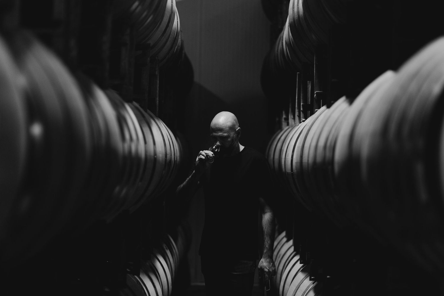 Peter Bartle tasting wine between rows of oak barrels in central otago wine cellar.