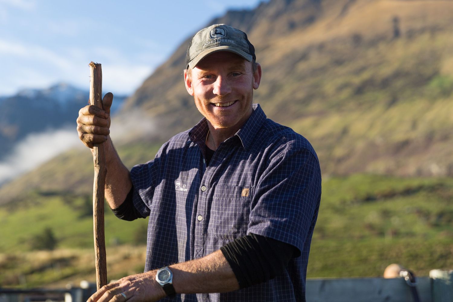 Smiling farmer holding wooden staff with snow-capped mountain backdrop on a sunny day in New Zealand.