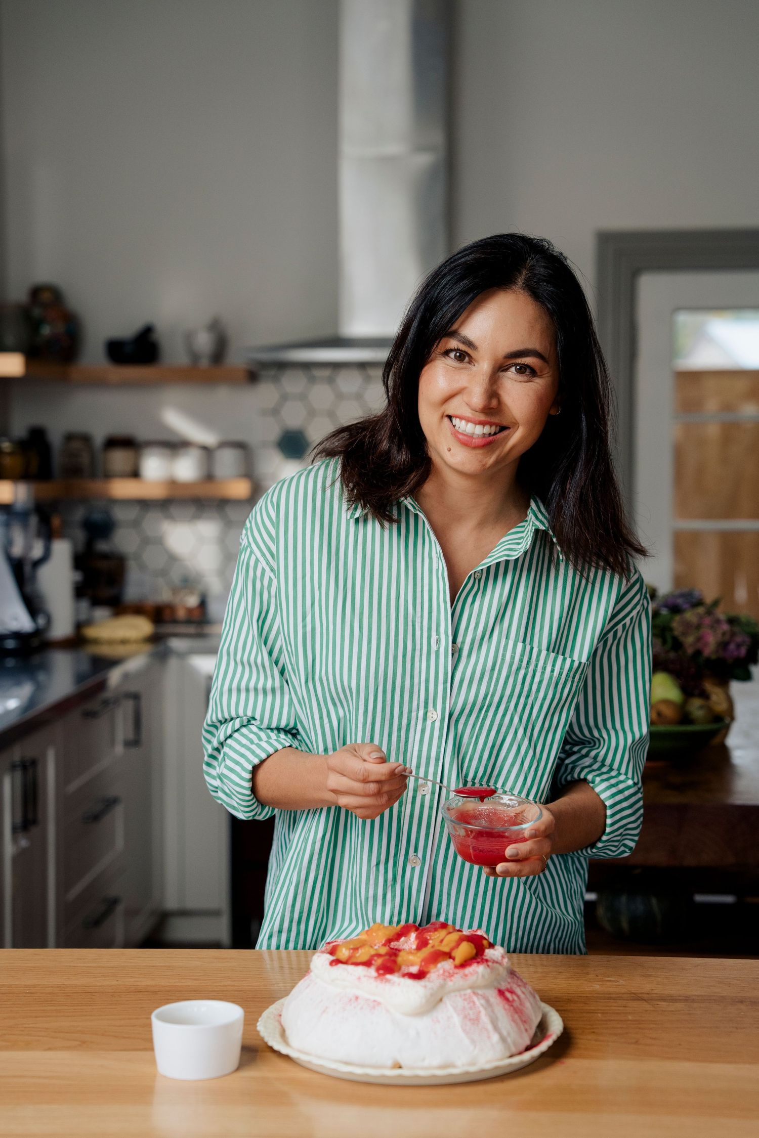 Smiling woman in striped shirt stands in modern kitchen beside a pavlova dessert.