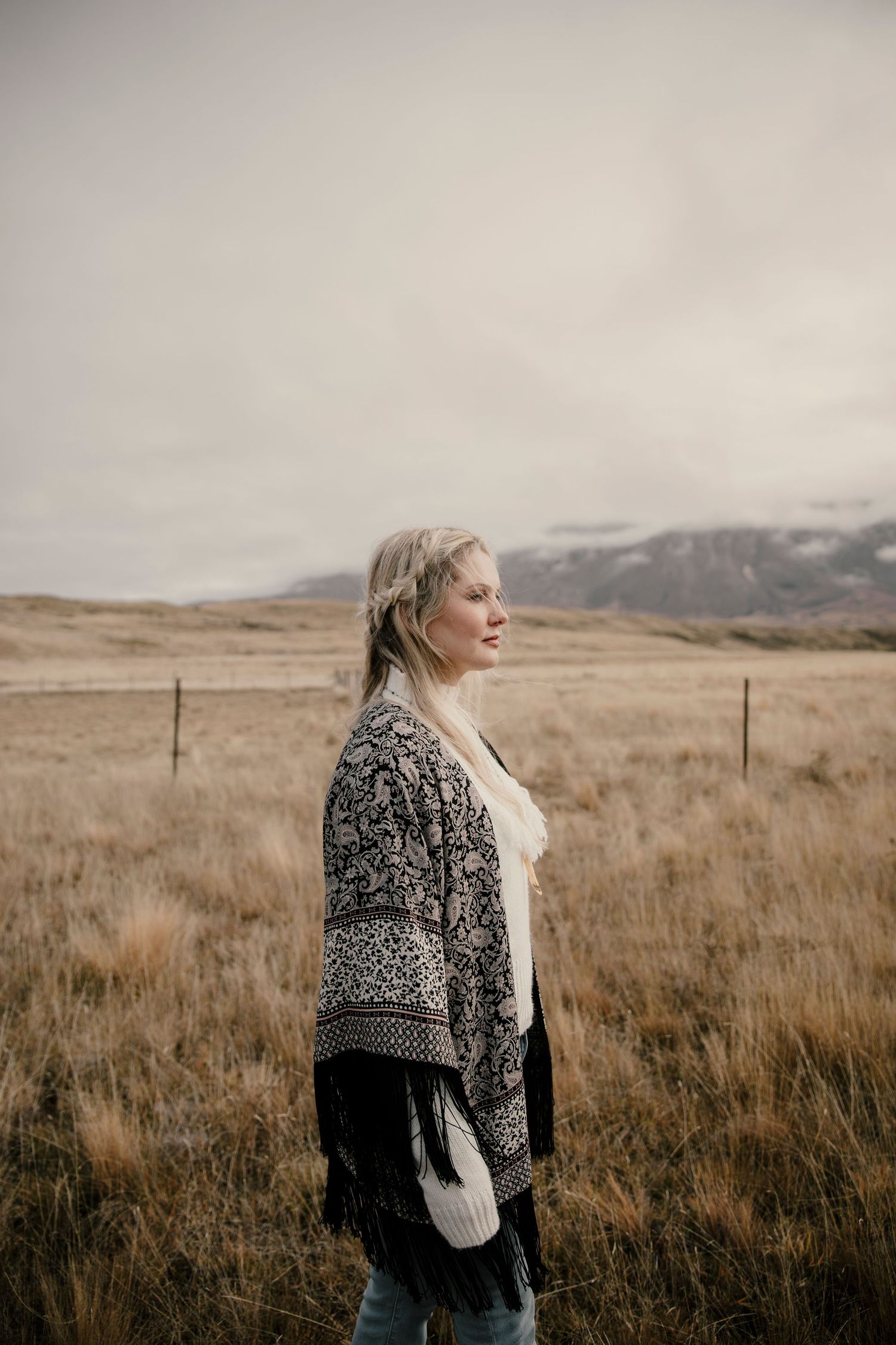 Blonde woman in patterned shawl standing in golden field with mountains behind.