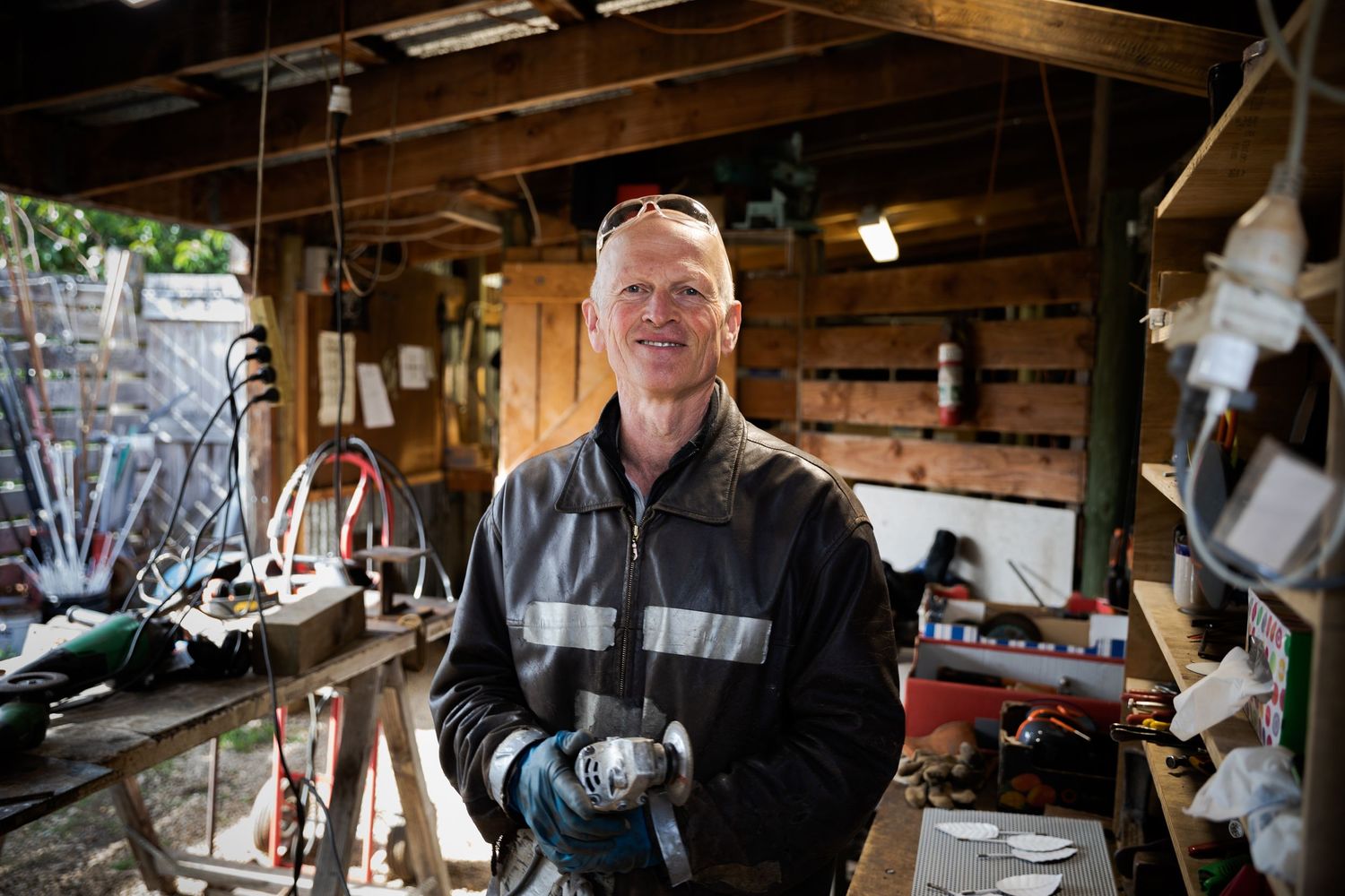 Smiling tradesman holding power tool in a well-equipped workshop garage filled with tools and equipment.