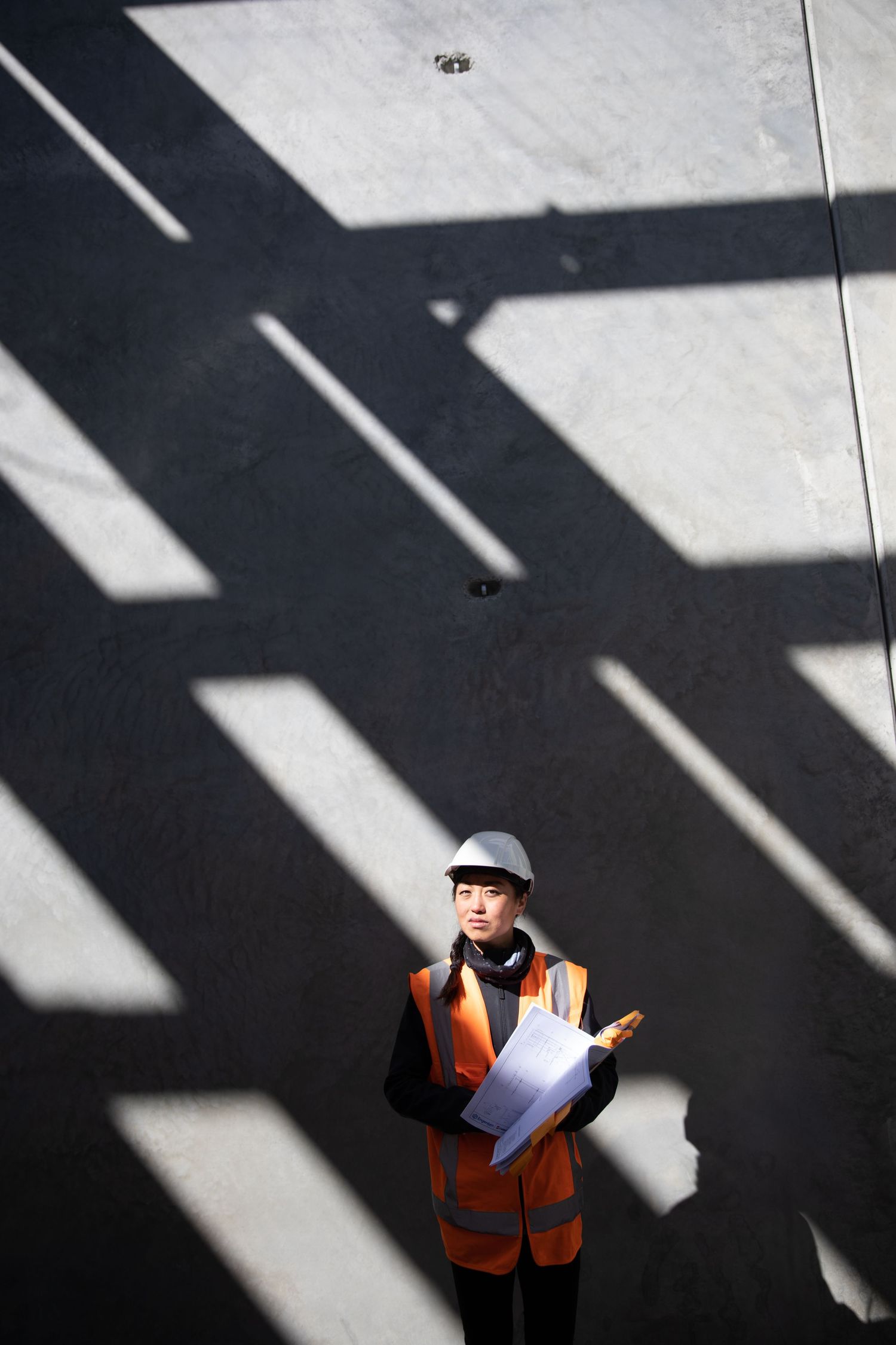 Aerial view of a female engineer in orange safety vest and white hard hat holding blueprints.