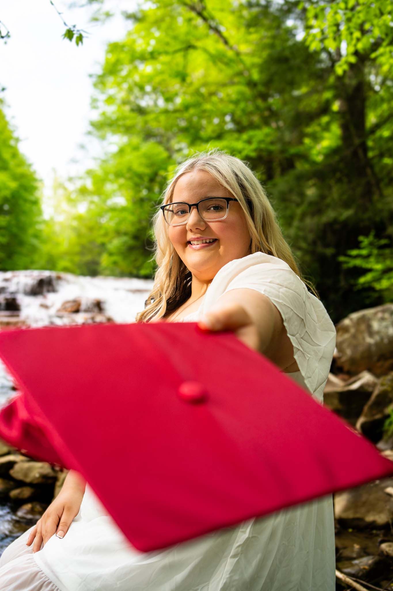 Smiling graduate holds red cap toward camera near forest waterfall.