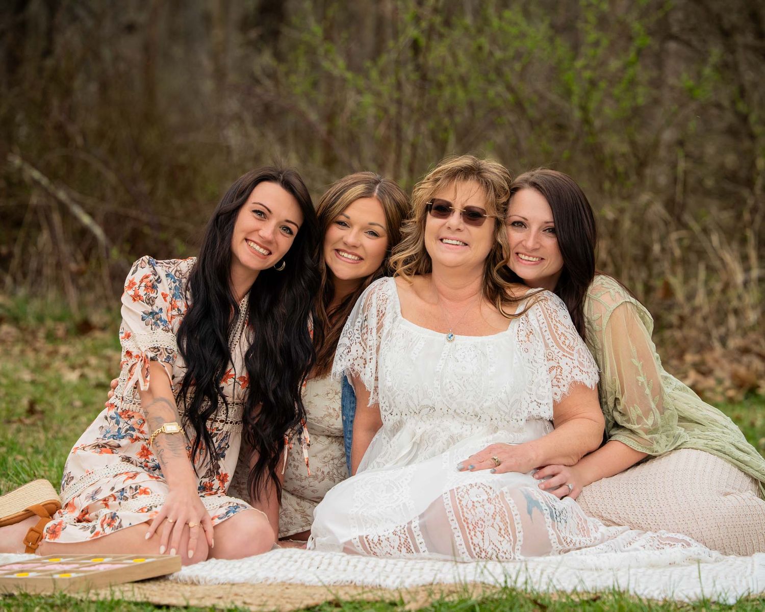 Four women smiling together while sitting on a blanket in a natural outdoor setting.