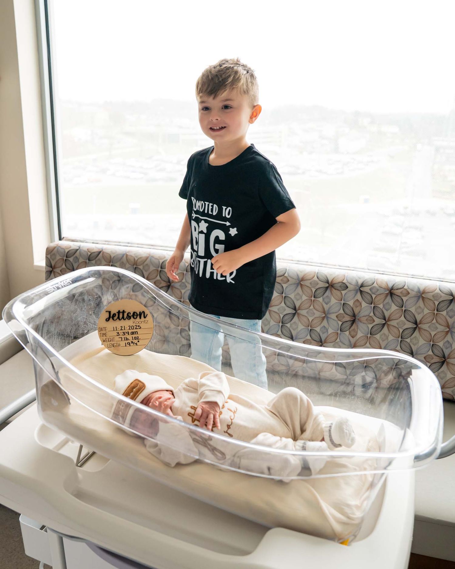 Young boy wearing a promoted to big brother shirt stands proudly beside a hospital bassinet with newborn baby.