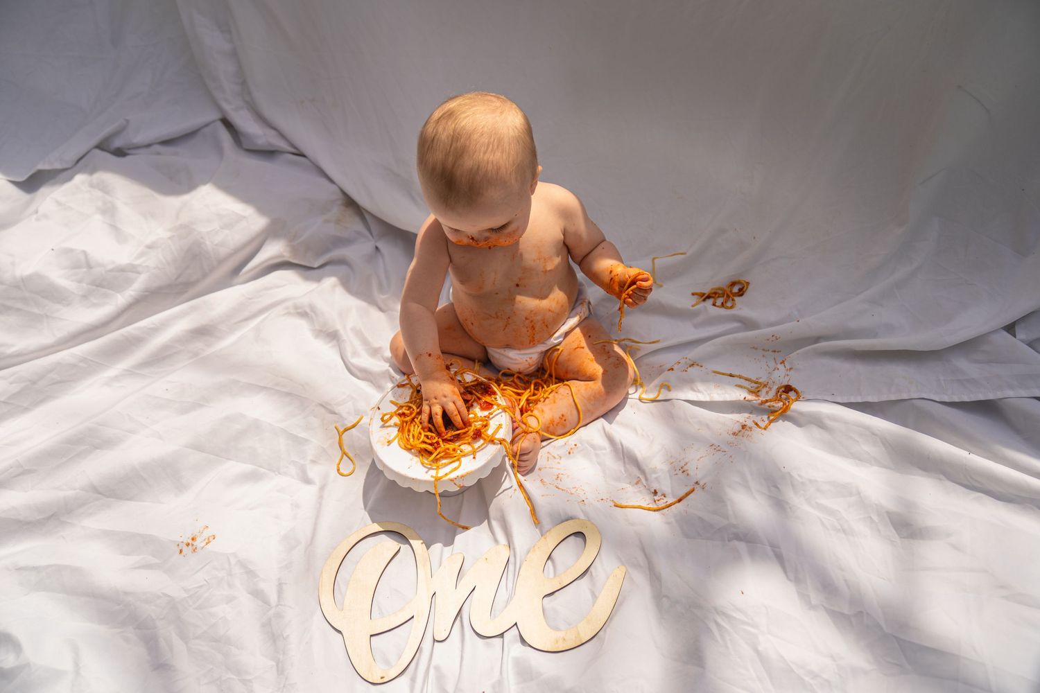 Baby in diaper celebrating first birthday smashing cake, with wooden 'one' sign on white fabric backdrop.