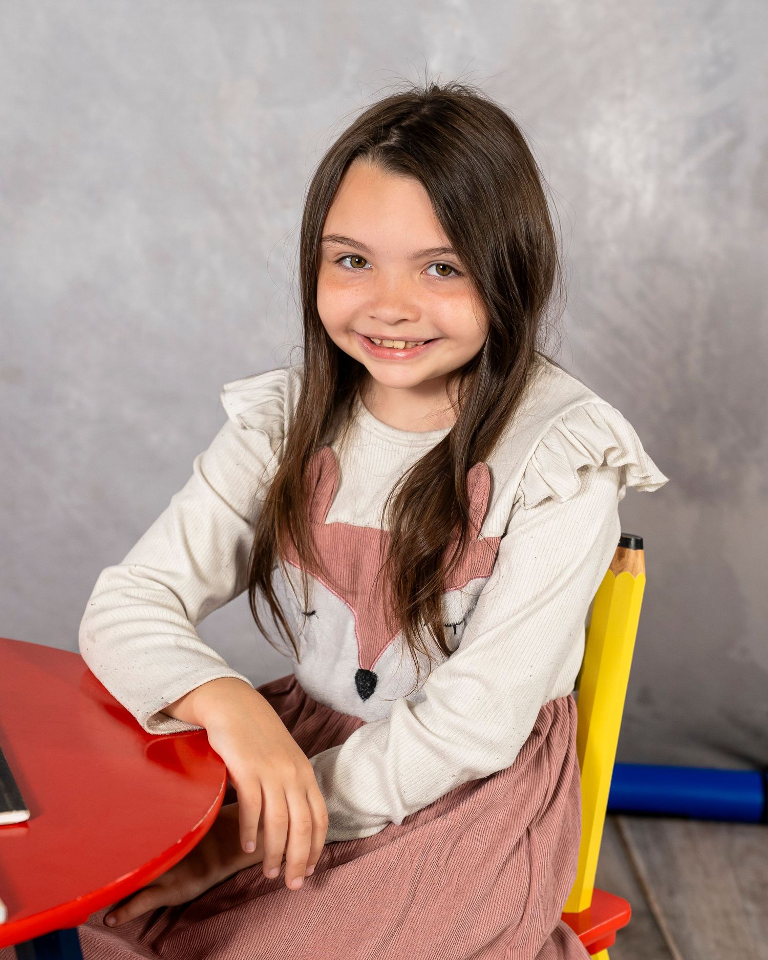 Young girl smiling in school portrait, wearing fox graphic shirt, seated at colorful desk.