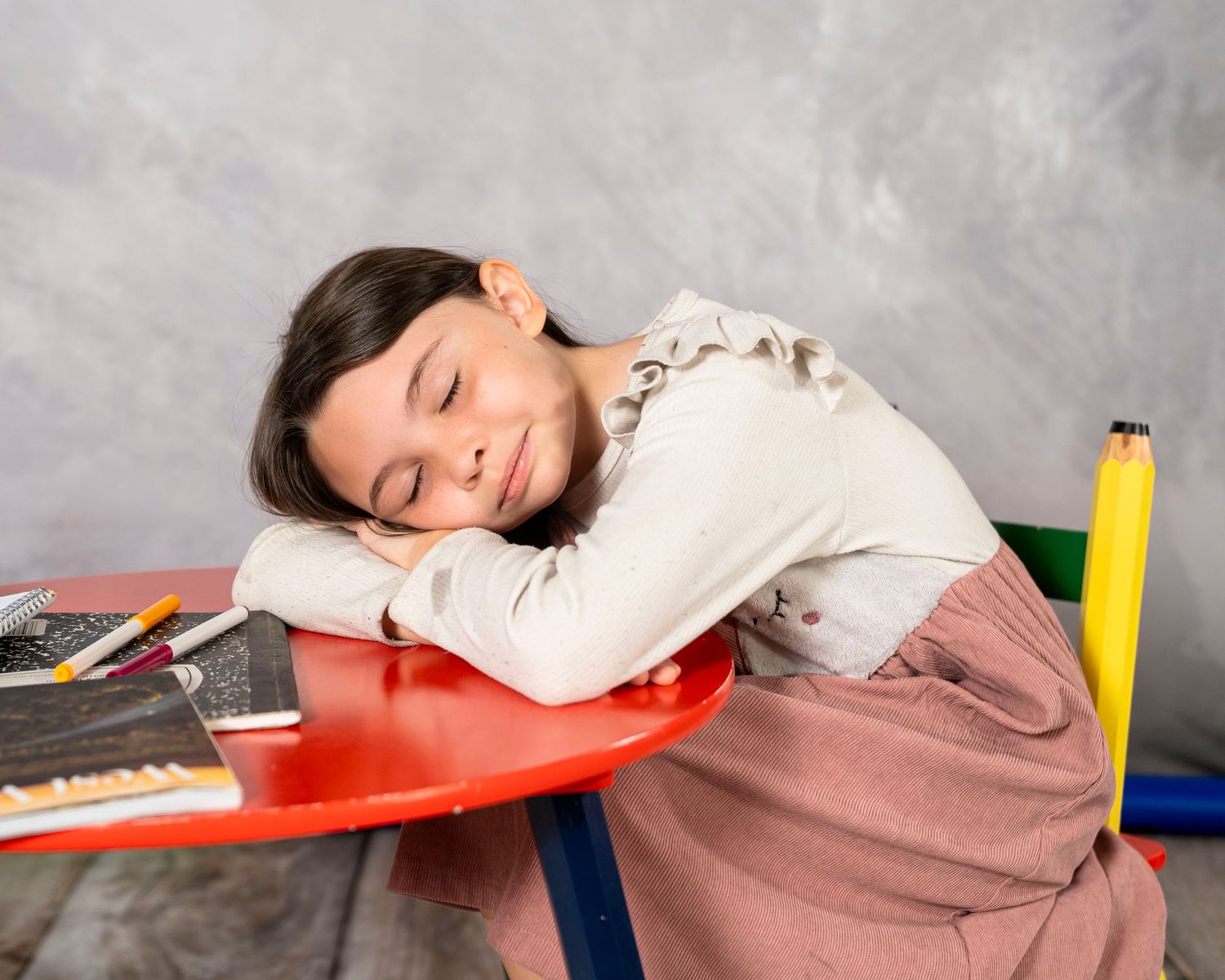 Young girl sleeping on a colorful desk with school supplies, resting her head on her arms during study time.
