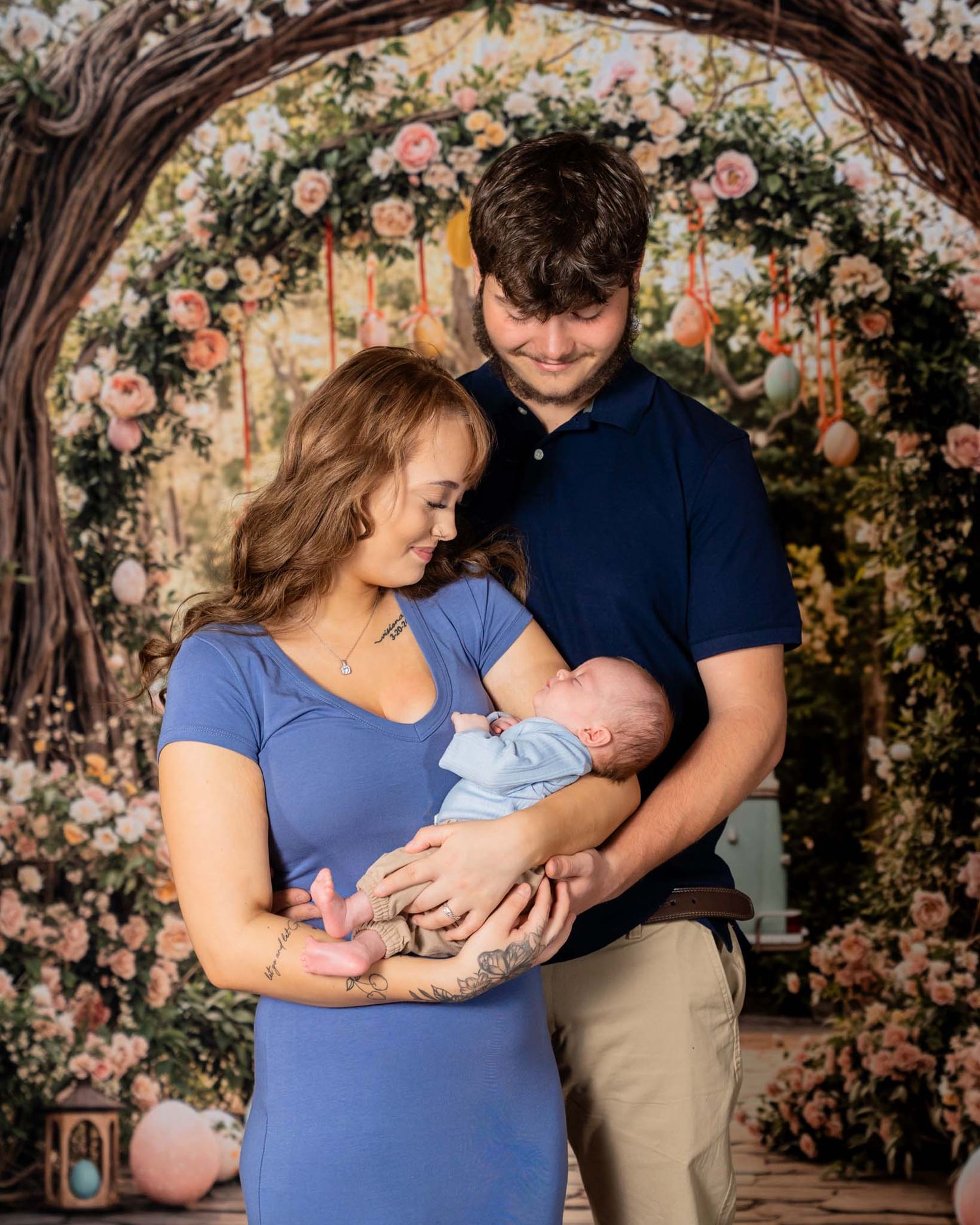 Couple in blue outfits holding newborn baby in front of floral arch backdrop in professional photo.