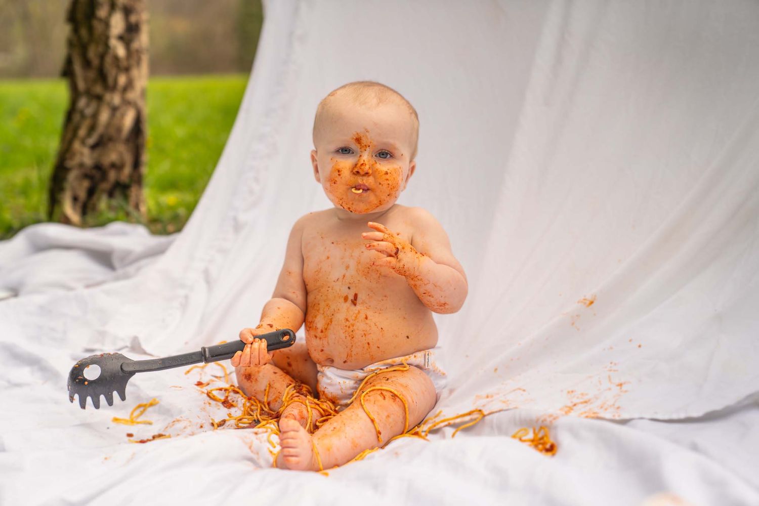 Messy baby covered in sauce sits outdoors on white blanket holding a pasta spoon.
