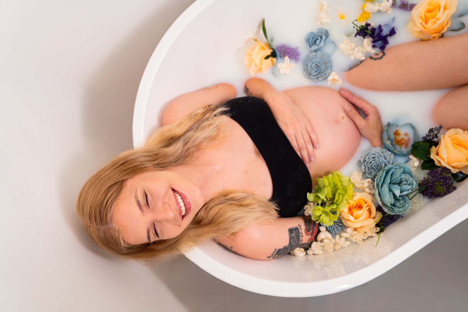 Smiling pregnant woman relaxing in floral milk bath with colorful flowers wearing black top.