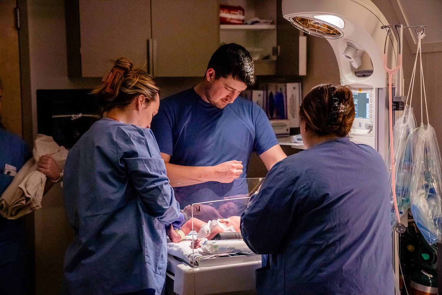 Medical team in blue scrubs examining newborn baby under warming lamp in hospital delivery room.