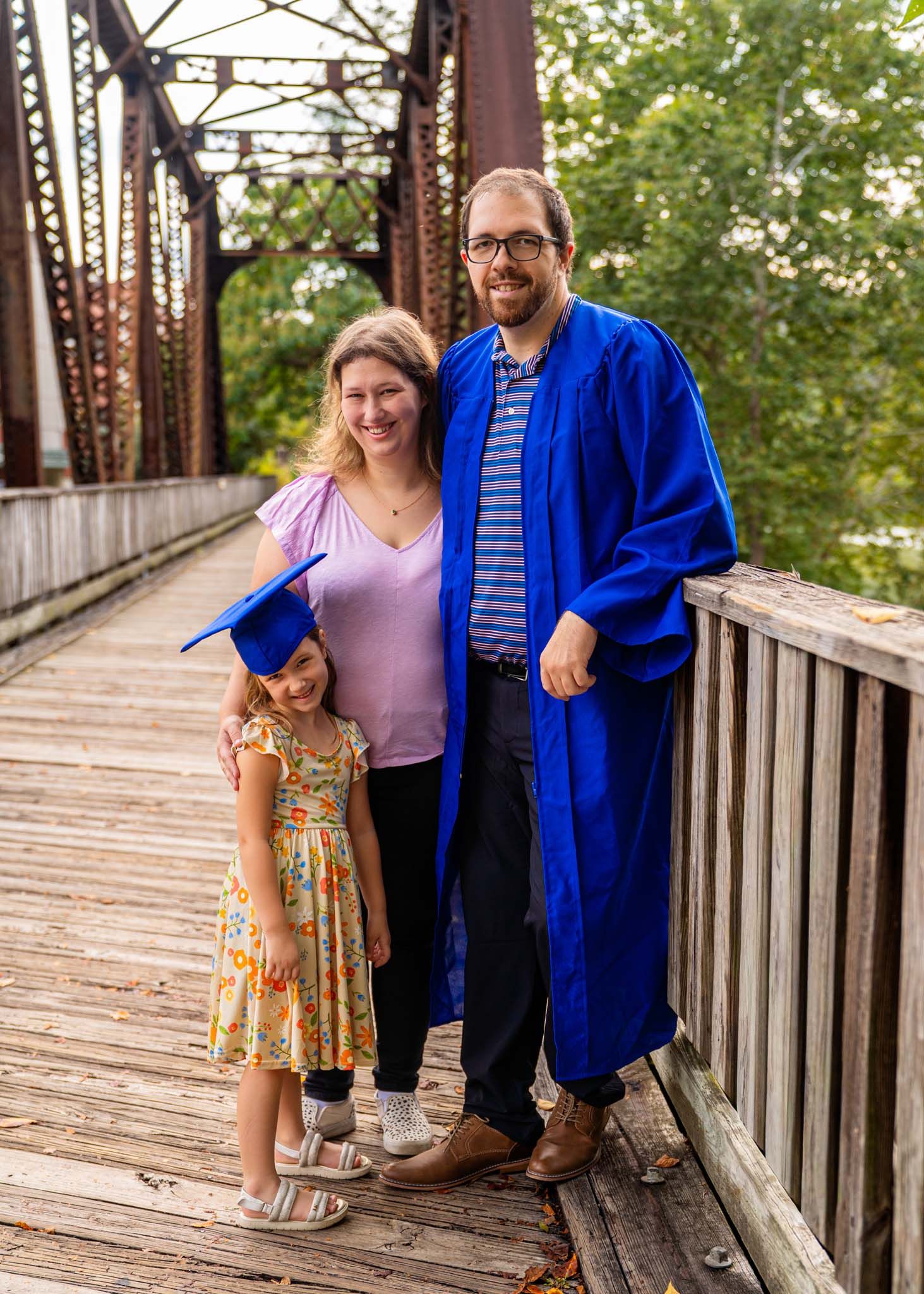 Family graduation photo on wooden bridge, man in blue cap and gown with woman and young girl.