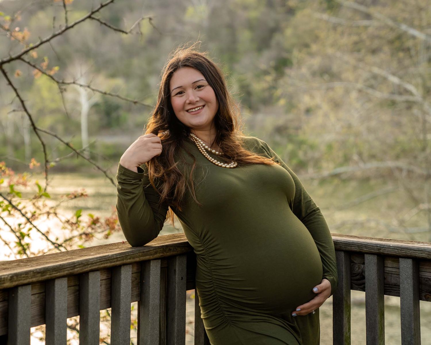 A smiling pregnant woman in an olive green dress leans on a wooden railing outdoors.