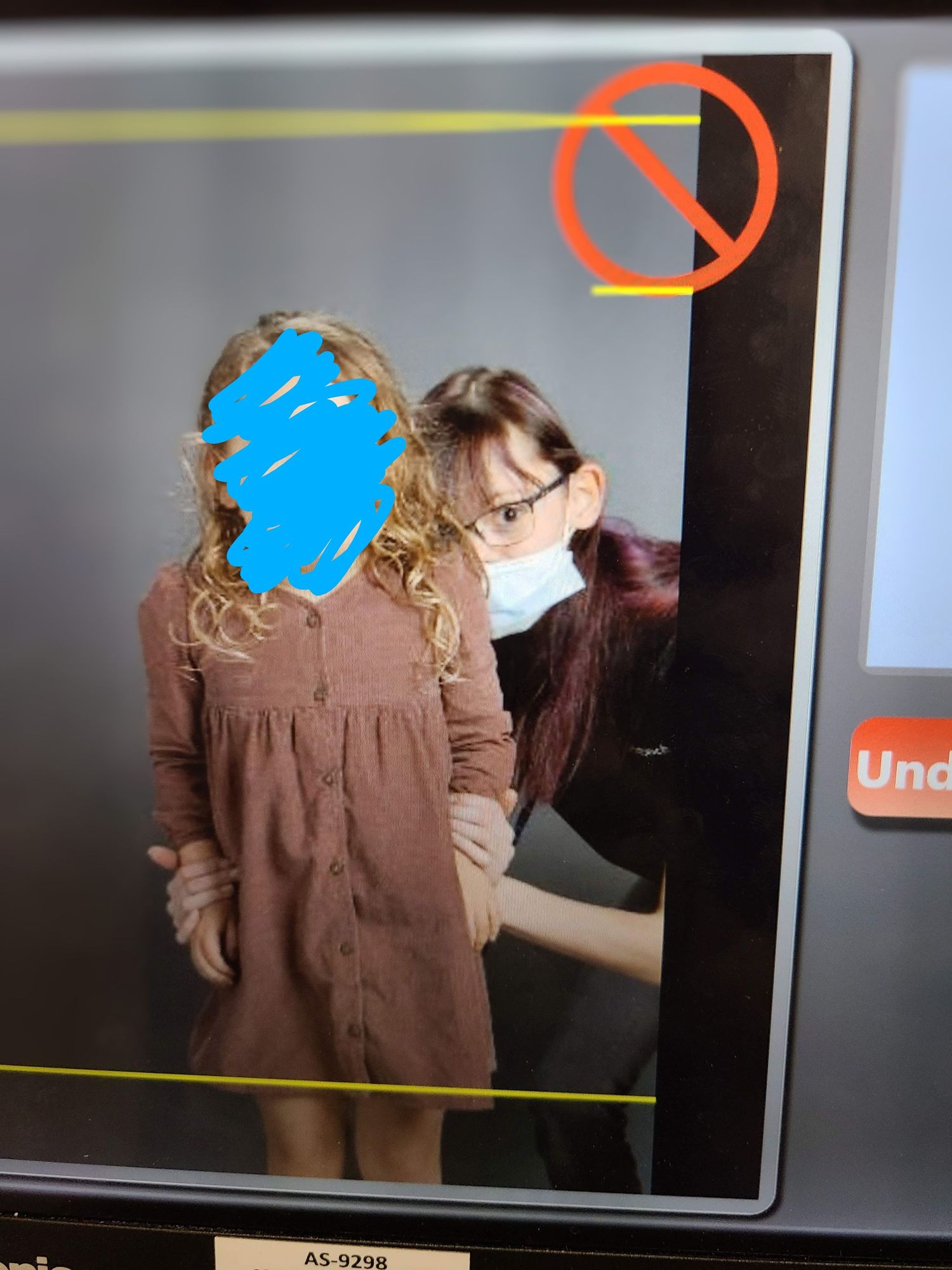 Woman wearing a face mask assists a young girl with curly hair in a brown button-up dress indoors.