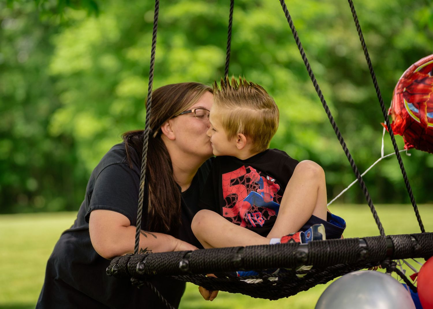 A mother kisses her young son on a swing in a lush green park, with a balloon visible nearby.