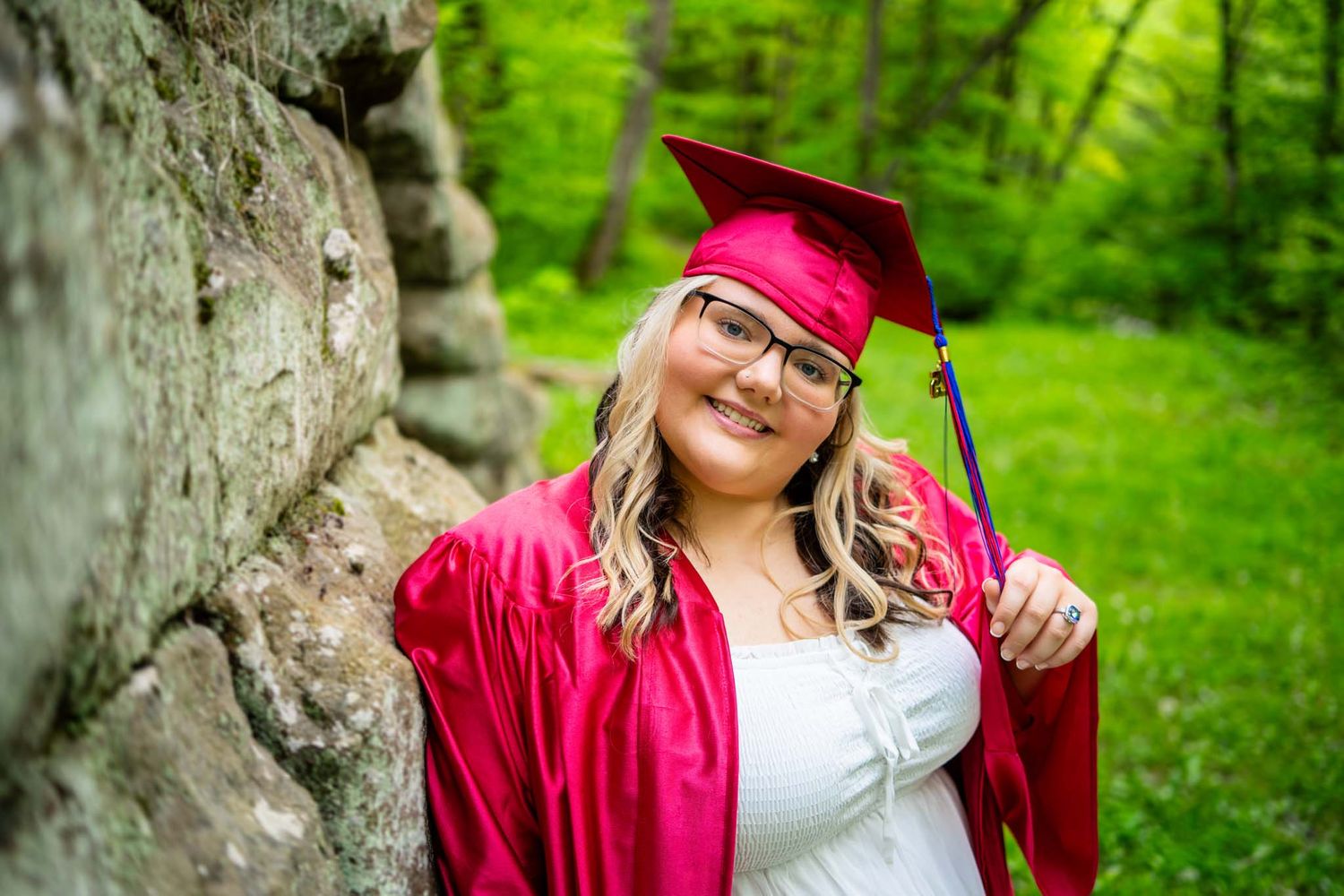 Smiling graduate in red cap and gown leans against stone wall outdoors.