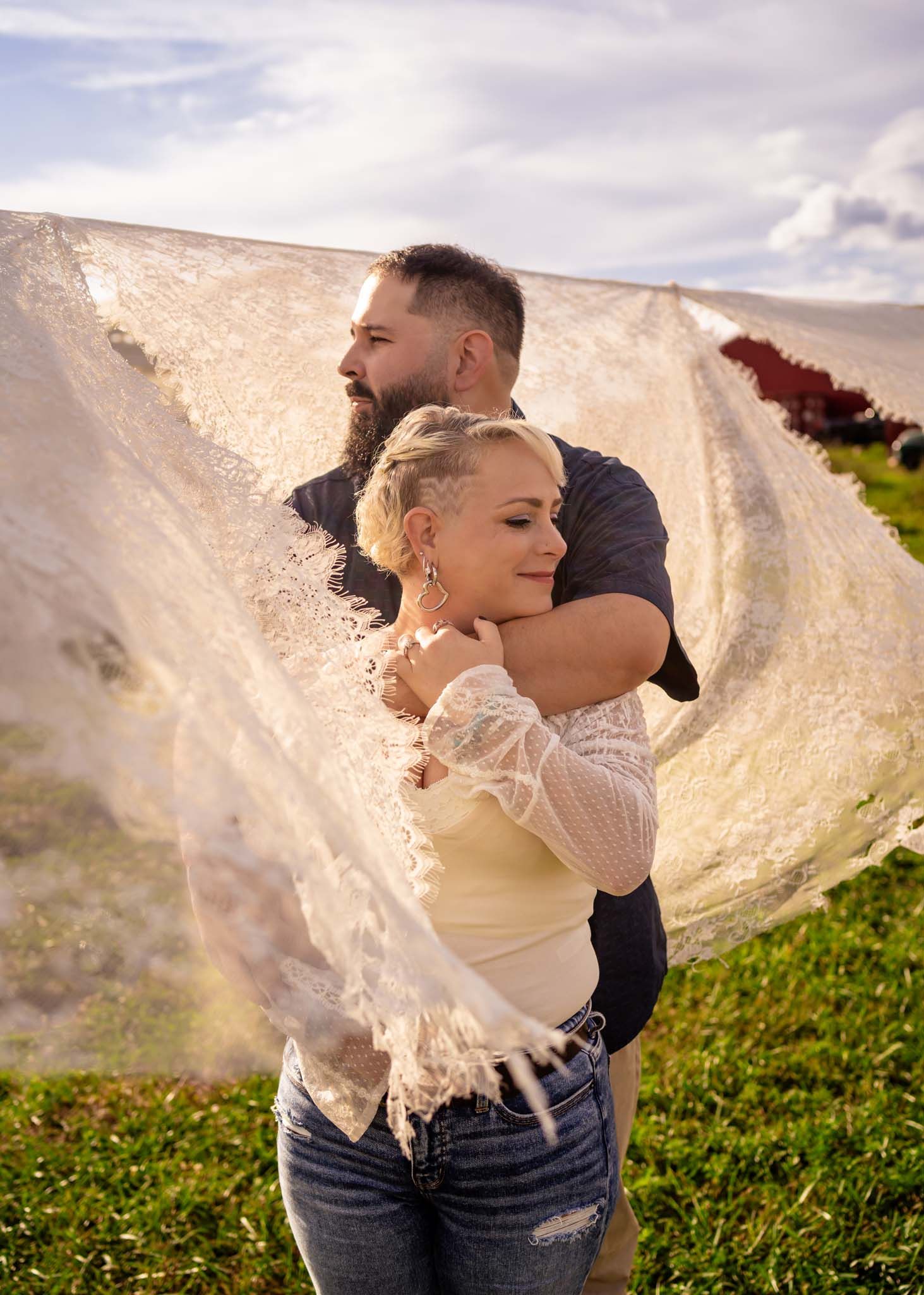 Man embraces woman from behind near billowing sheer fabric in golden outdoor light.
