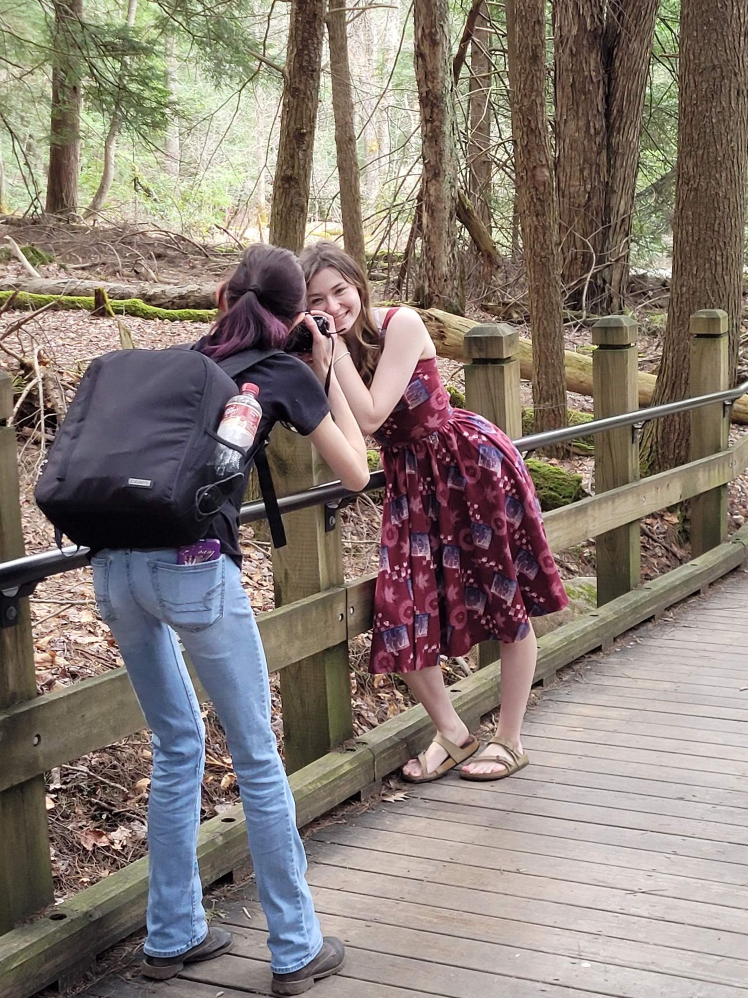 A photographer takes a portrait of a smiling woman in a floral dress on a wooden bridge in a lush forest.