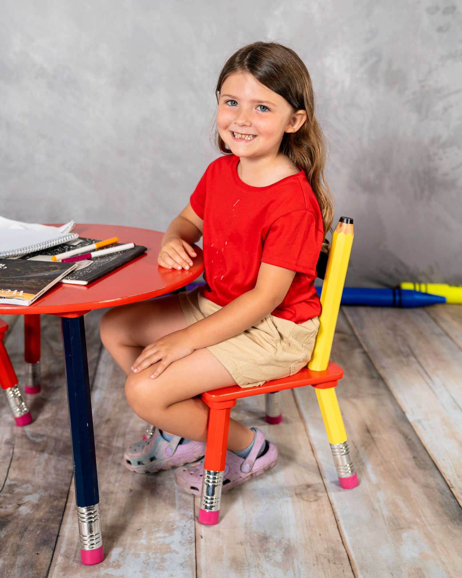 Smiling girl in red shirt sits at colorful pencil-shaped kids table and chair set on wood floor.