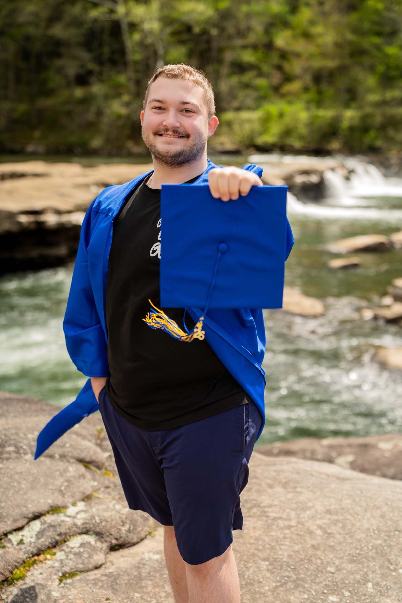 Smiling male graduate in blue gown holding cap toward camera near waterfall outdoors.