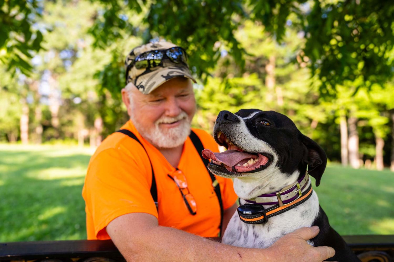 Smiling older man in orange shirt affectionately hugs a black and white dog outdoors in a park.