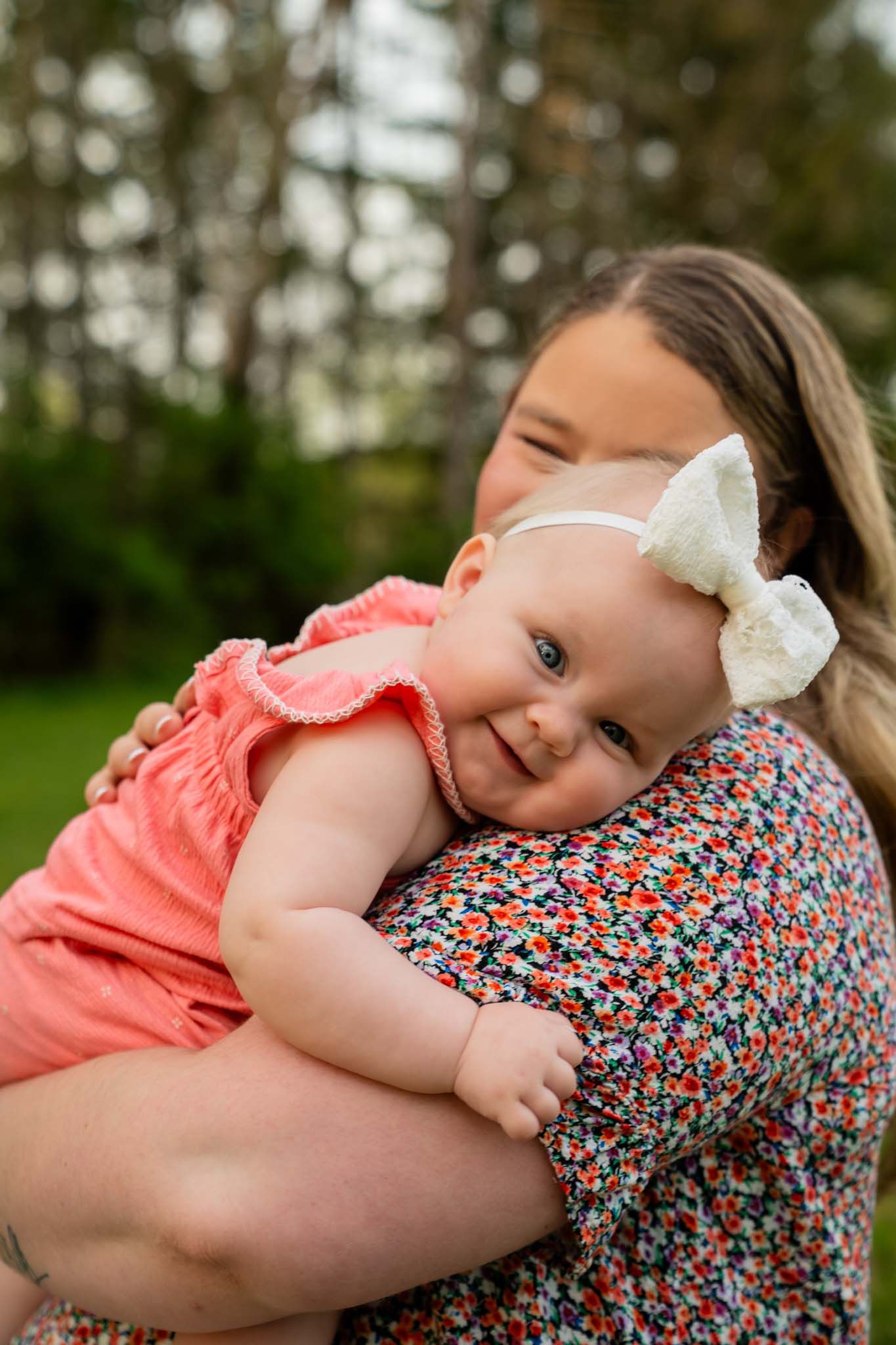 Mother holding smiling baby girl in pink dress with white bow headband outdoors.