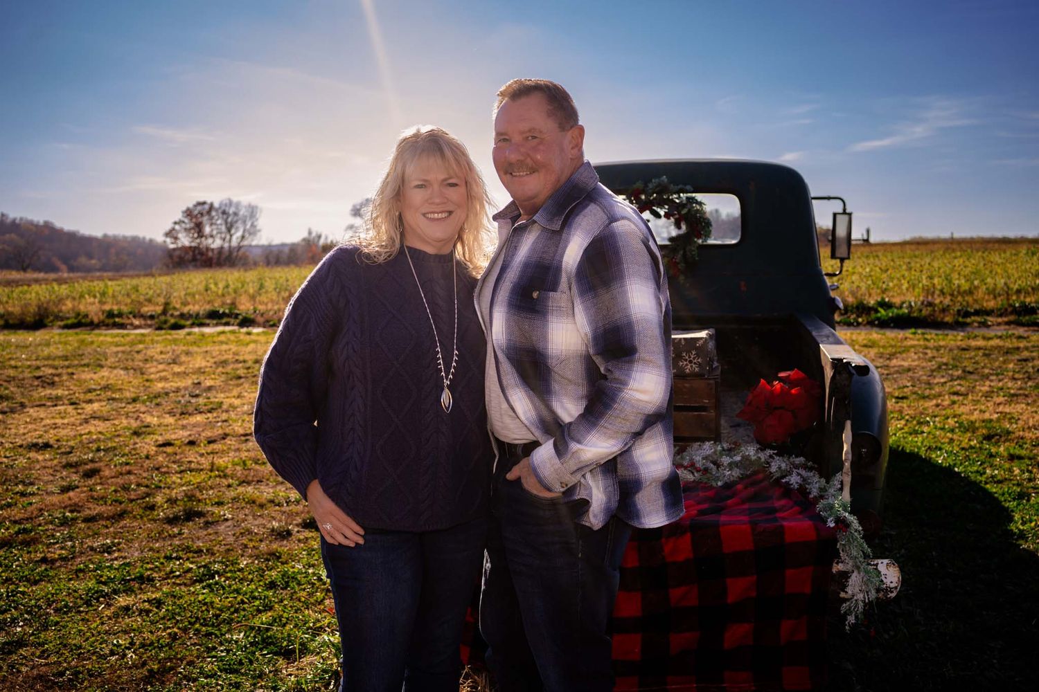Smiling couple standing beside a vintage Christmas-decorated truck in a sunny open farm field.